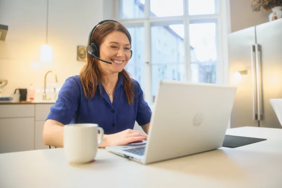 Eine Frau mit Headset sitzt lächelnd vor einem Laptop, daneben steht eine Tasse