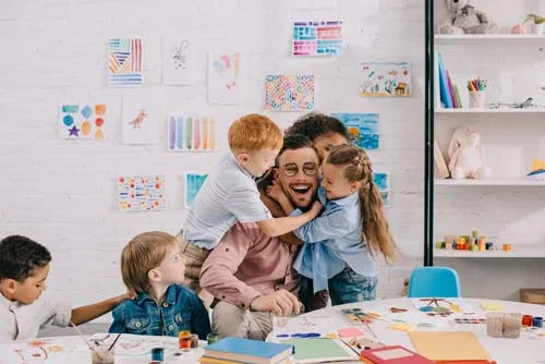 Erzieher im Klassenzimmer wird von lachenden Kindern umarmt.